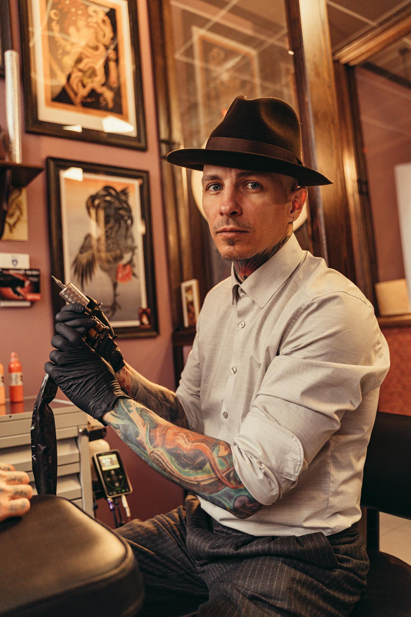 Caucasian tattoo artist sitting indoors, holding tattoo machine, wearing fedora and gloves.