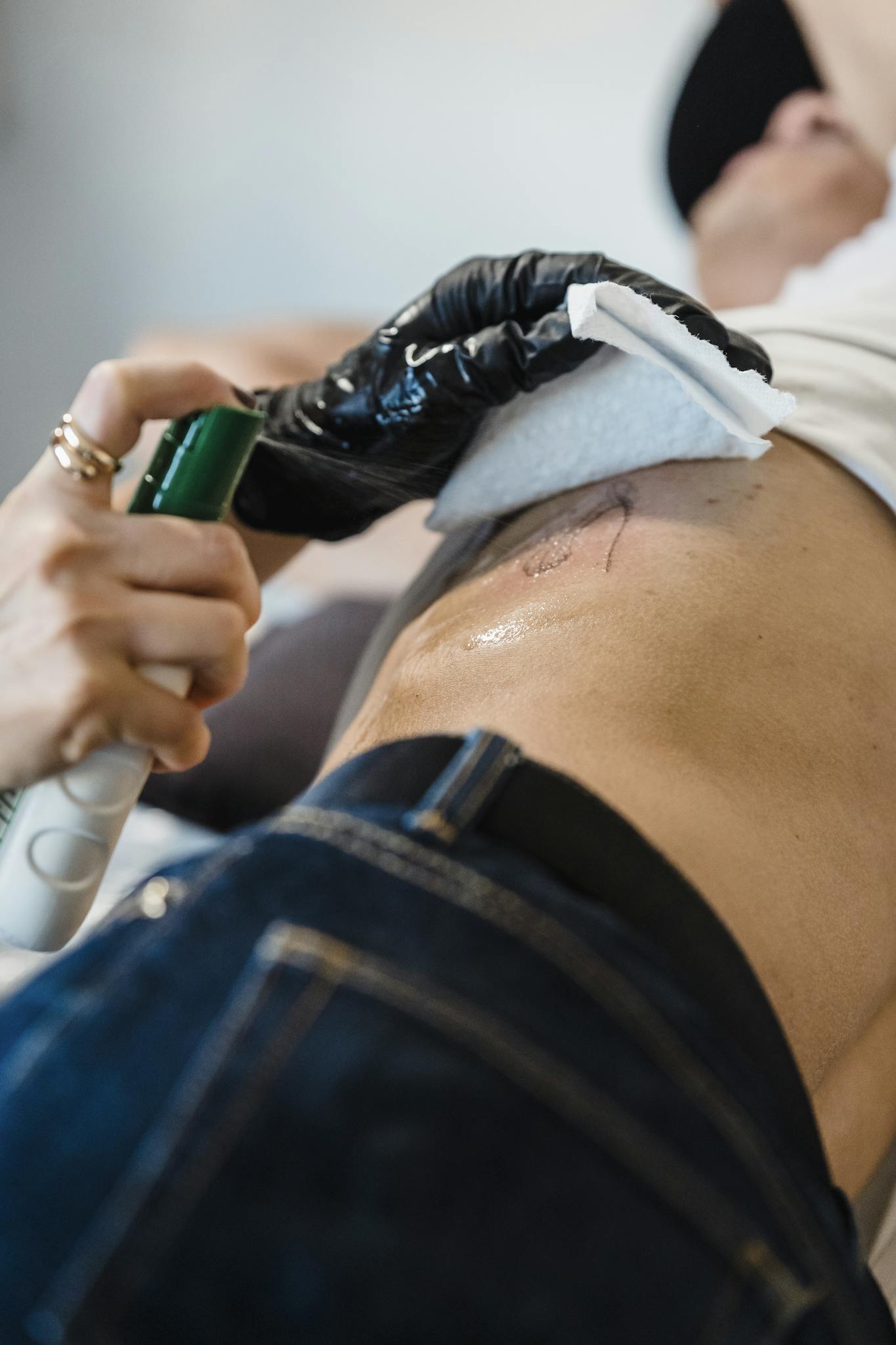 Crop unrecognizable female tattoo master in sterile gloves spraying antiseptic on body of anonymous client in light salon